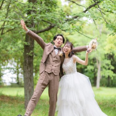 新緑の木立の中で両手を広げて足を上げ、はしゃぐツイードの新郎とウェディングドレスの花嫁。元気な前撮りポーズ