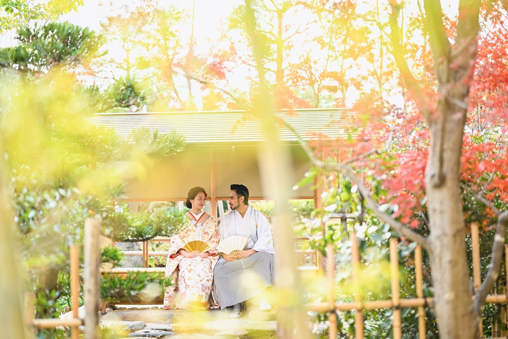 紅葉に囲まれた東屋のベンチに並んで座る色打掛の花嫁と紋付袴の新郎。竹垣越しに撮る和装ロケーションフォト