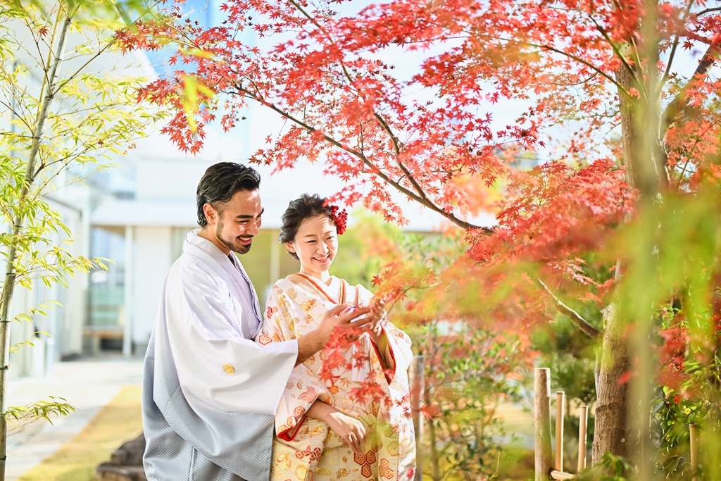 藤田邸跡公園の紅葉の下で見つめ合う色打掛の花嫁と白い羽織袴の新郎。色づく秋の和装前撮り