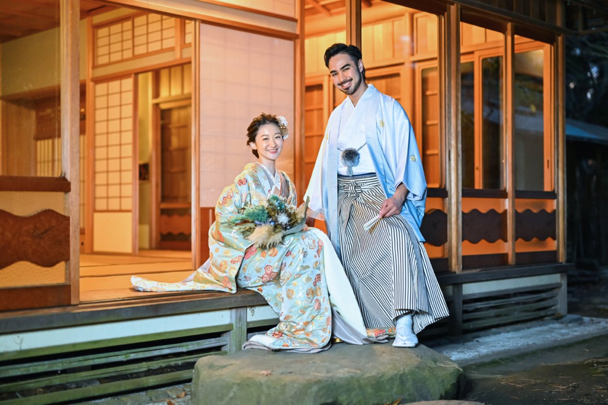 むらさき亭の縁側に座る花嫁と傍らに立つ新郎の和装ツーショット