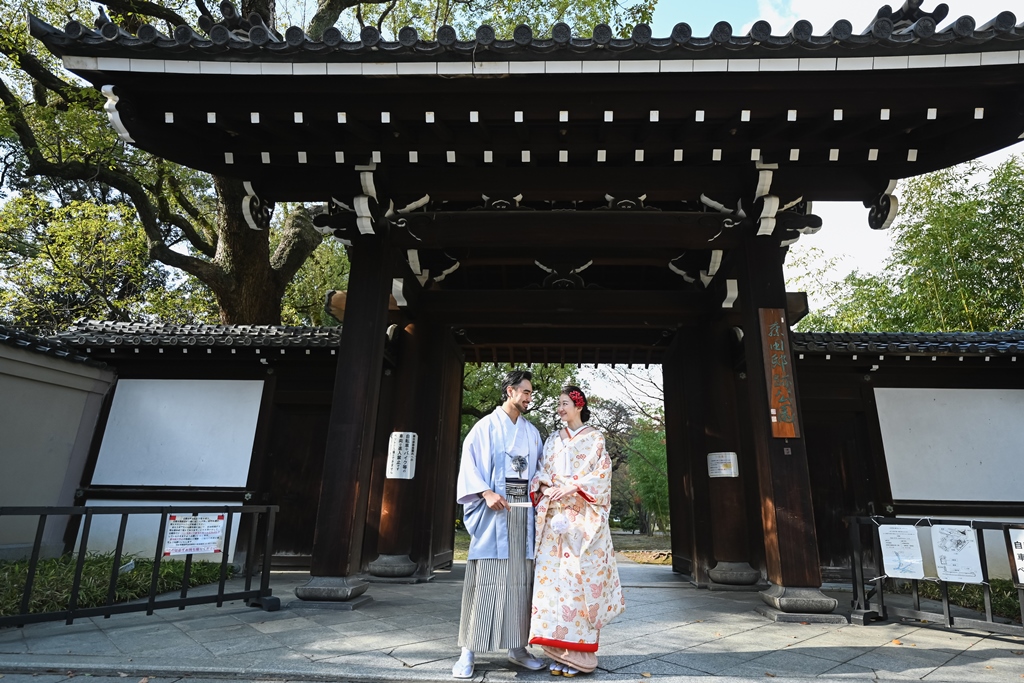 黒い木造の大きな門の前に並ぶ紋付袴の新郎と色打掛の花嫁。歴史を感じる門で撮る和装ロケーションフォト