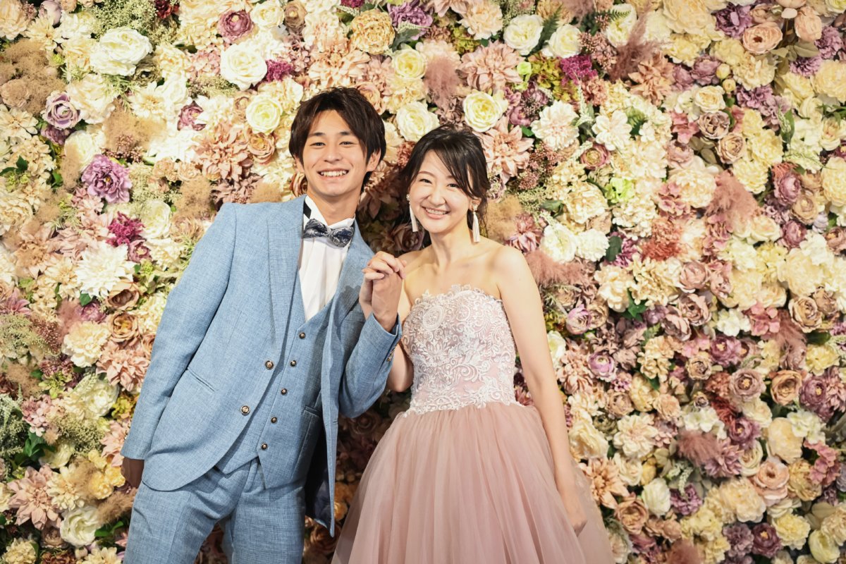 くすみピンクのカラードレスと水色スーツで並ぶ新郎新婦｜春色フラワーウォールのフォトウェディング