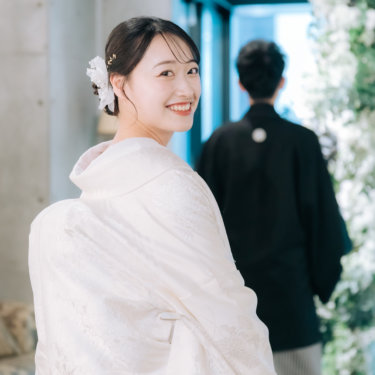 グリーンの装飾の前で振り向いて満面の笑みを見せる白無垢の花嫁と、背を向けた新郎。ファーストミートの和装前撮り