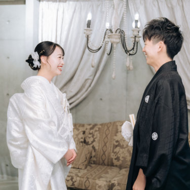 シャンデリアのあるセットで向き合い、顔を見合わせて笑う紋付の新郎と白無垢の花嫁。ファーストミートの和装前撮り