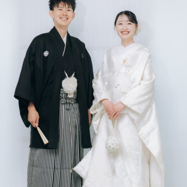 白い背景に並んで立ち、笑顔を見せる紋付袴の新郎と白無垢の花嫁。心斎橋のスタジオで撮る和装前撮り