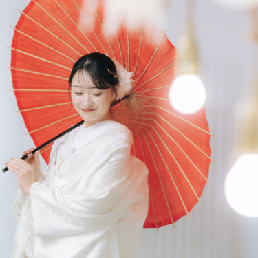電球の飾りを前ぼかしに、赤い和傘を肩にかけてうつむいてほほえむ白無垢の花嫁のソロカット。和装前撮り