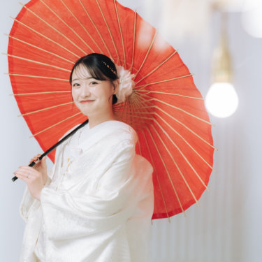 電球の飾りを前ぼかしに、赤い和傘を肩にかけてこちらにほほえむ白無垢の花嫁のソロカット。スタジオ和装前撮り