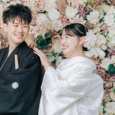 フラワーウォールの前で新郎の肩に手を添え、笑い合う紋付の新郎と白無垢の花嫁のアップ。スタジオ和装前撮り