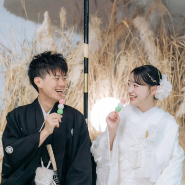 ススキのセットで三色団子を手に、顔を見合わせて笑う紋付の新郎と白無垢の花嫁。遊び心のある和装前撮り