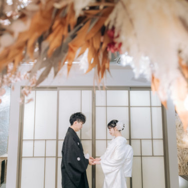ドライフラワーの飾りを前ぼかしに、障子の前で手を取り合う紋付の新郎と白無垢の花嫁のスタジオ和装前撮り