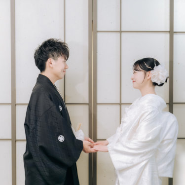 障子の前で手を取り合い、見つめ合ってほほえむ紋付の新郎と白無垢の花嫁。心斎橋のスタジオで撮る和装前撮り