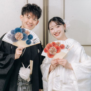 '夫''妻'の花飾りの扇子を胸元に持ち、満面の笑みを見せる紋付の新郎と白無垢の花嫁の和装前撮り