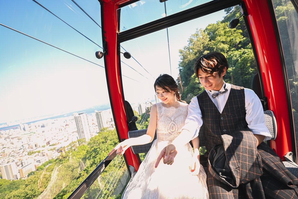神戸布引ロープウェイの赤いゴンドラの中で、窓の外の街並みを指さすレースドレスの花嫁とベストの新郎の前撮り