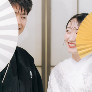 白と金の扇子で顔を半分隠し、目元だけで笑い合う紋付の新郎と白無垢の花嫁。遊び心のある和装前撮り