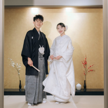 金色の壁の床の間の前に並んで立ち、ほほえむ紋付袴の新郎と白無垢の花嫁。梅の枝を飾った和装前撮り