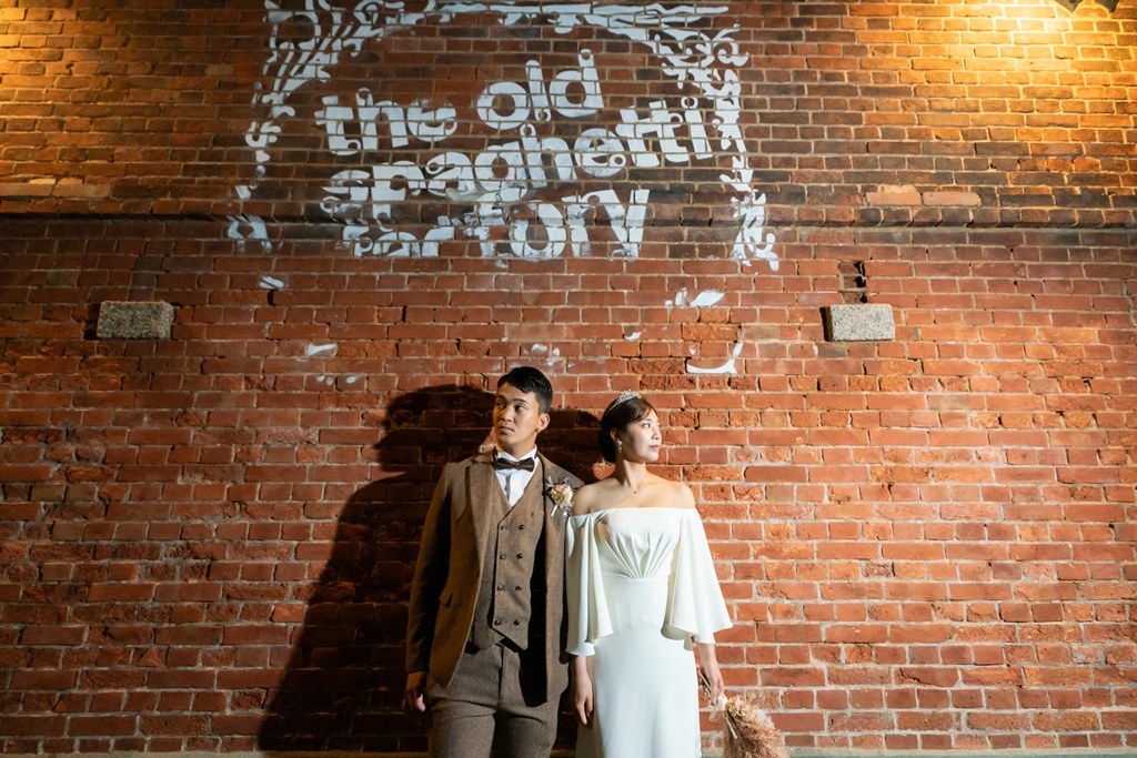 赤レンガ倉庫の壁を背景に並ぶ、ブラウンスーツの新郎とオフショルダードレスの新婦のロケーション前撮り