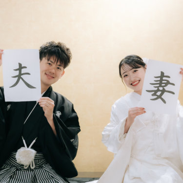 '夫''妻'の書を顔の横に掲げて笑う紋付の新郎と白無垢の花嫁。金色の壁の和室で撮る和装前撮り