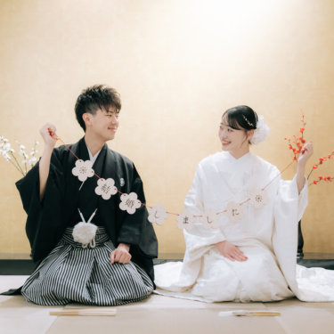 金色の壁の床の間の前で梅の花のガーランドを広げ、顔を見合わせる紋付袴の新郎と白無垢の花嫁の和装前撮り