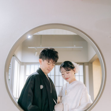 丸窓の中で寄り添い、手元を見てほほえむ紋付の新郎と白無垢の花嫁。円窓を額縁にしたスタジオ和装前撮り