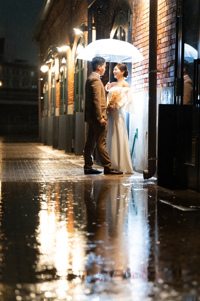 雨の夜の神戸ハーバーランド、赤レンガの壁の前で傘をさして見つめ合う新郎新婦と濡れた路面のリフレクションが美しい前撮り