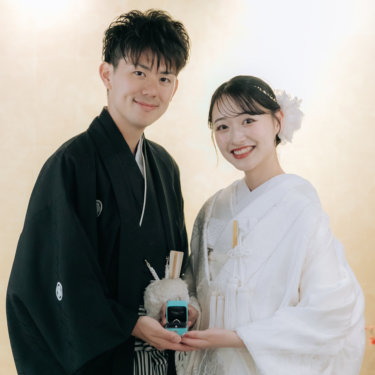 指輪の箱を一緒に持ち、こちらに笑顔を見せる紋付の新郎と白無垢の花嫁。金色の壁を背にした和装前撮り