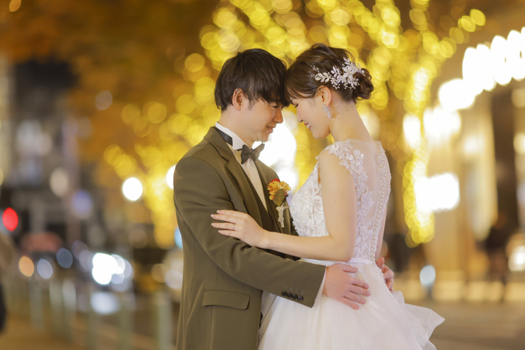 冬の夜、街路樹のイルミネーションの光に包まれて額を寄せ合う新郎新婦のナイトロケーションフォト