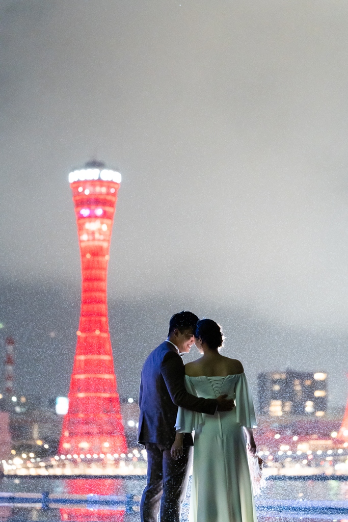 雨の夜、赤く灯る神戸ポートタワーを望む海辺で寄り添う新郎新婦のナイトロケーションフォト