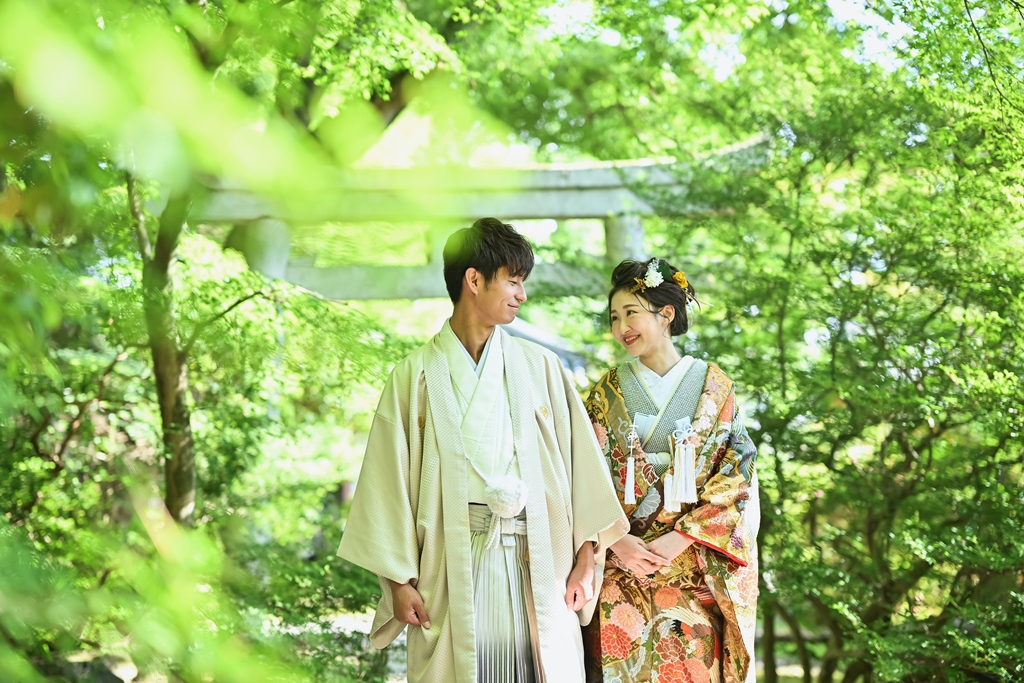 木々の緑の中にたたずむ鳥居の前で手をつないで見つめ合う、色打掛の花嫁と羽織袴の新郎のフォトウェディング