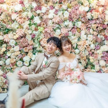フラワーウォールの前に座るベージュスーツの新郎と白ドレスの花嫁｜家族やペットと叶えるフォトウェディング