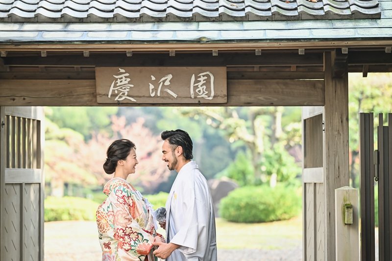 「慶沢園」の扁額が掲げられた門の下で見つめ合う、水色の紋付羽織の新郎と花柄着物の花嫁