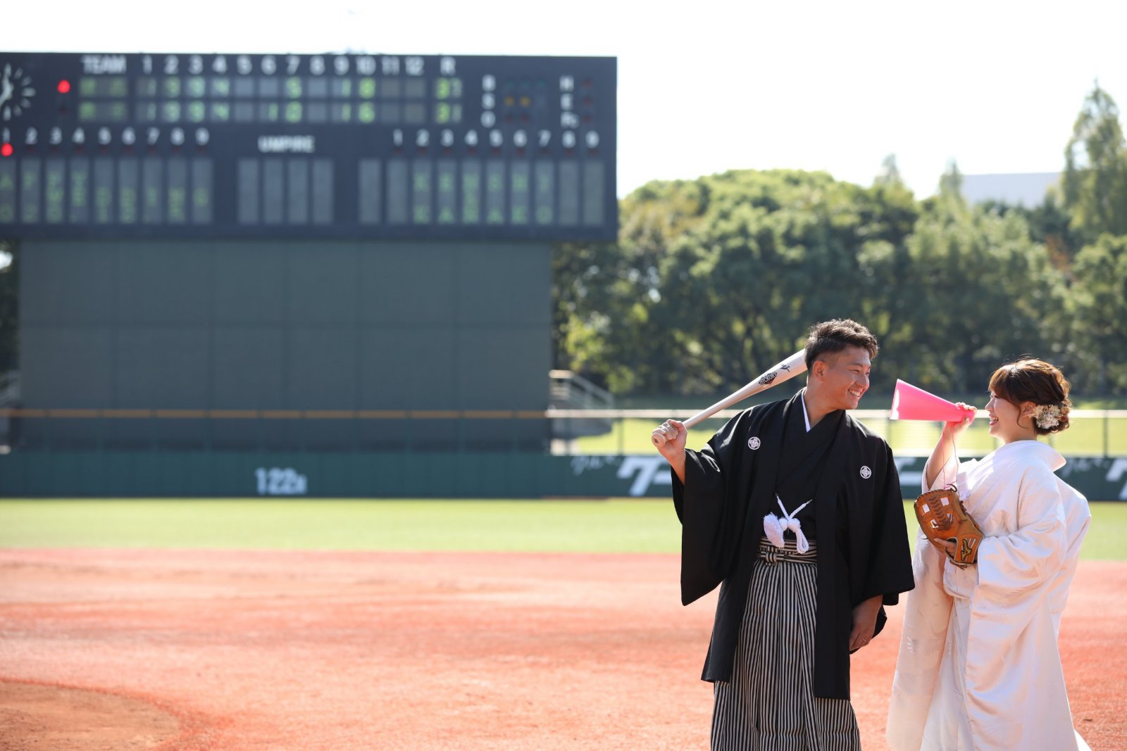 野球場のスコアボードを背景に、バットを担ぐ黒紋付袴の新郎とグローブとピンクのメガホンを持つ白無垢姿の花嫁