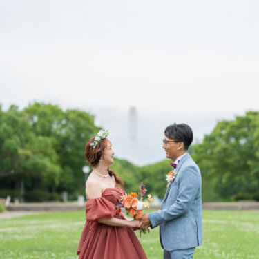 芝生広場で向かい合い、ブーケを手に笑い合う新郎新婦。カラードレスのロケーションフォトウェディング