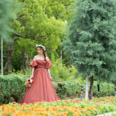 オレンジの花壇のそばを歩く、花冠とテラコッタのカラードレスの花嫁。花いっぱいの公園での前撮り