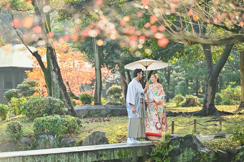 紅葉のもみじ越しに石橋の上で見つめ合う、水色の紋付羽織の新郎と白い和傘の下で花柄着物の花嫁