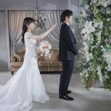 白いカーテンとシャンデリアのスタジオで、ブーケを持って手を挙げるウェディングドレスの花嫁と黒いタキシードの新郎。心斎橋店のスタジオで撮る洋装前撮り