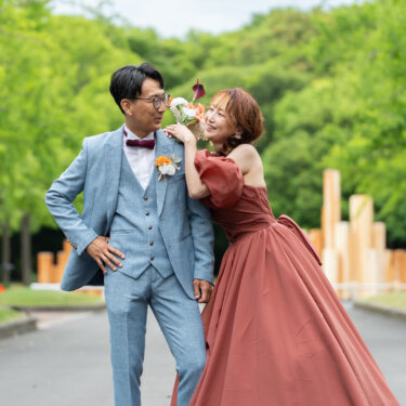 並木道でブーケを新郎の肩に寄せ、楽しそうに笑う花嫁とライトブルースーツの新郎。大阪での洋装ロケーションフォト