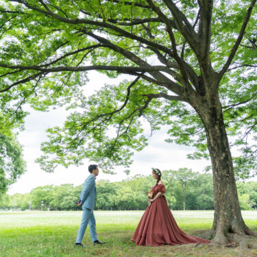 大木の下に佇む花嫁のもとへ歩み寄る新郎。芝生と新緑が広がる公園でのフォトウェディング
