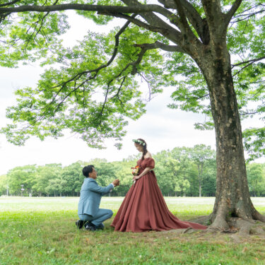 大きな木の下でプロポーズする新郎と、見下ろして微笑むテラコッタドレスの花嫁。新緑の公園ロケーションフォト