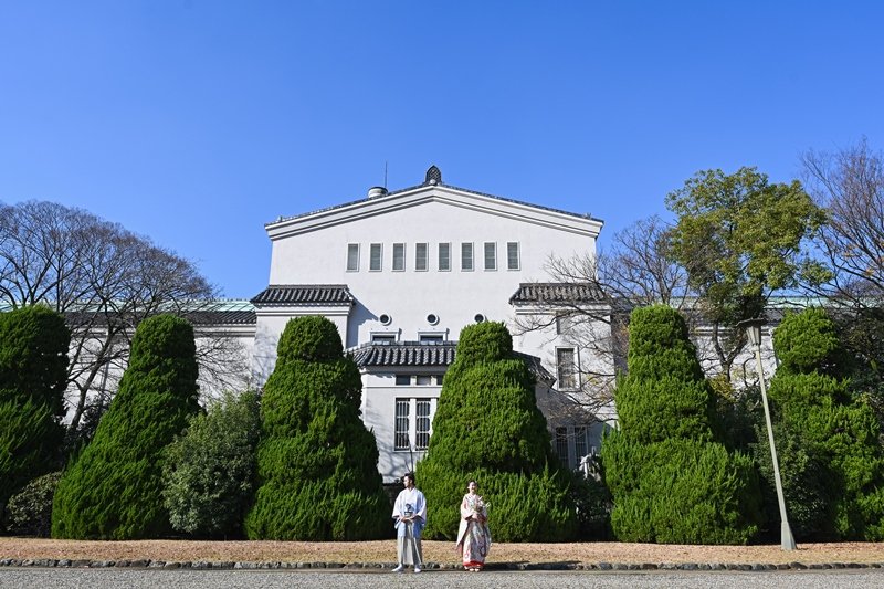 青空と白亜の洋館を背景に並んで立つ、灰色の紋付羽織の新郎と朱赤の着物を着た花嫁（遠景）
