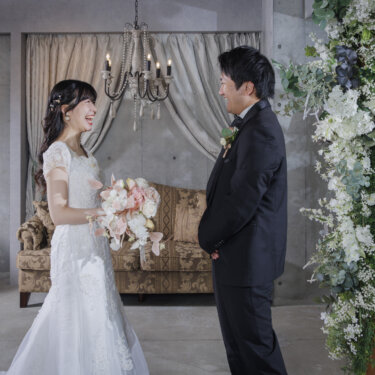 白いカーテンとシャンデリアのスタジオで、ブーケを持って向かい合うウェディングドレスの花嫁と黒いタキシードの新郎。心斎橋店のスタジオで撮る洋装前撮り