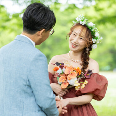 花冠をつけたテラコッタドレスの花嫁が、新郎と手を取り合って笑顔を見せる新緑の公園での前撮り
