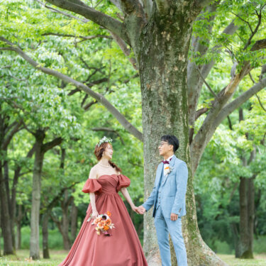 大木の下で手をつなぎ、見つめ合う花冠の花嫁とライトブルースーツの新郎。新緑の公園での前撮り