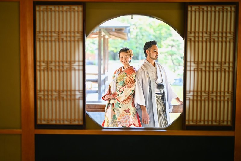 円窓越しに背中合わせで立つ、水色の紋付羽織の新郎と花柄着物の花嫁