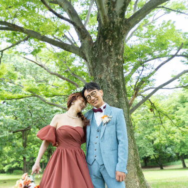 大きな木の幹を背に、頭を寄せ合って目を閉じる新郎新婦。ブーケを携えたカラードレスのロケーションフォト