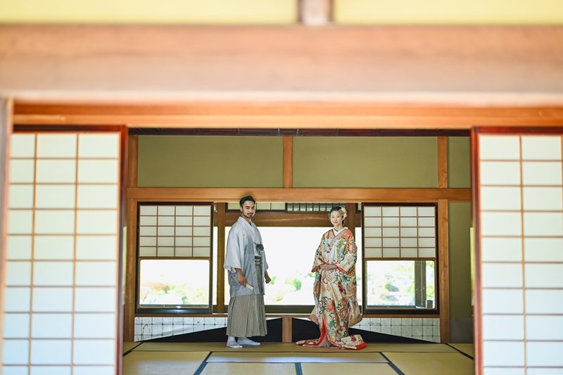 障子を開けた広間で並んで立つ、水色の紋付羽織の新郎と花柄着物の花嫁