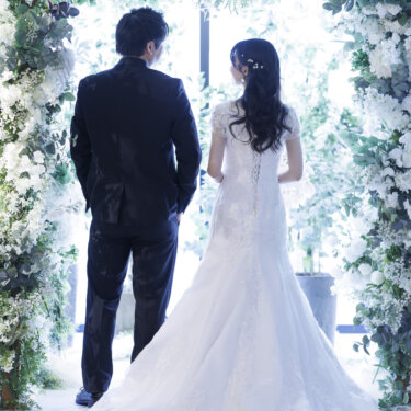 白いフラワーアーチの下、窓の光を見つめて並んで立つ新郎新婦の後ろ姿。ロングトレーンが広がるウェディングフォト