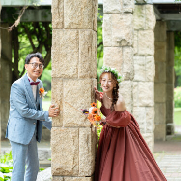 石の柱が並ぶ回廊で、柱の陰からのぞく水色のスーツの新郎とワインレッドのカラードレスの花嫁。大泉緑地の回廊で撮る洋装前撮りの遊び心のあるカット