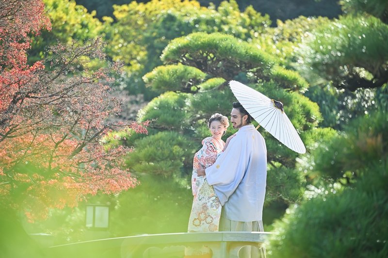 松の木々に囲まれ、白い和傘の下で寄り添う水色の紋付羽織の新郎と花柄着物の花嫁