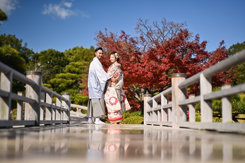 真っ赤な紅葉を背景に、木橋の上で寄り添う水色の紋付羽織の新郎と花柄着物の花嫁