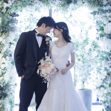 白い花と緑のフラワーアーチの下で、額を寄せ合う新郎新婦。光あふれるスタジオでの洋装前撮り